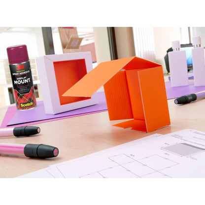 Une table avec un modèle en carton orange, des marqueurs, un spray adhésif 3M™ Scotch® DisplayMount™ (400 ml, 1 boîte/paquet) de 3M Deutschland GmbH, des boîtes blanches et des dessins techniques dans un espace de travail intérieur lumineux pour un usage professionnel.