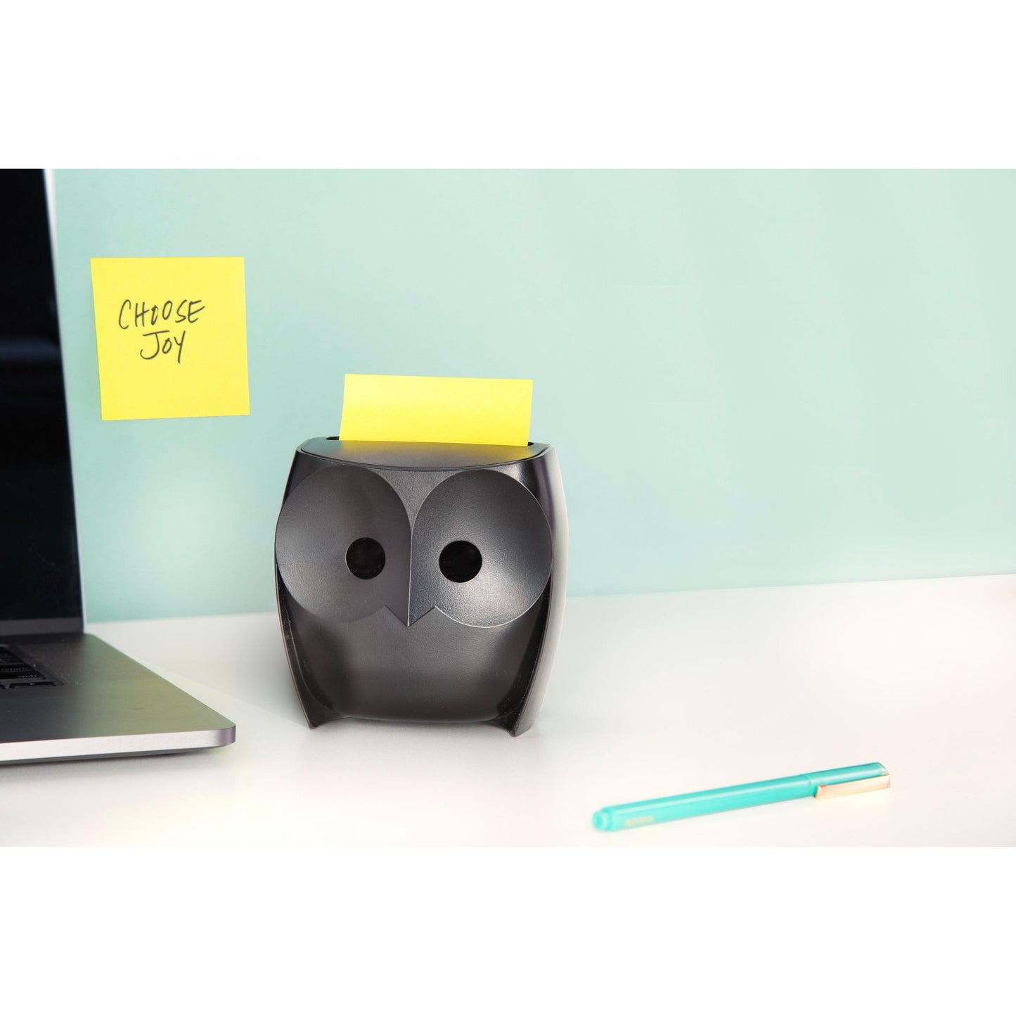 Ein schwarzer Post-it® Z-Zettelspender "Eule" (3M Deutschland GmbH) mit gelben Super Sticky Z-Zetteln steht auf einem weißen Schreibtisch, daneben ein Laptop und ein blauer Stift. An der Wand hängt ein "CHOOSE JOY"-Zettel vor hellblauem Hintergrund.
