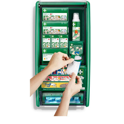 Une personne tient un bandage de gel pour brûlures devant la station de premiers secours & brûlures Cederroth (1 paquet) d'Orkla Wound Care AB, une station de premiers secours verte avec du matériel pour brûlures, des bandages, des bouteilles et des instructions.