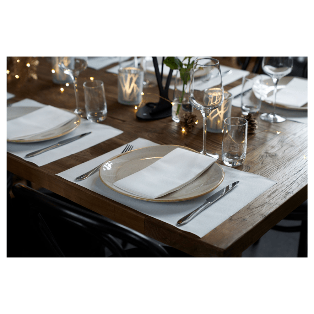 Une table à manger en bois dressée avec des sets de table blancs, des assiettes, des verres et des couverts. Les serviettes pliées Duni Dunilin® 48 x 48 cm blanches de Duni GmbH ajoutent du charme. Des bougies allumées et de petites lumières décoratives renforcent l'atmosphère conviviale.