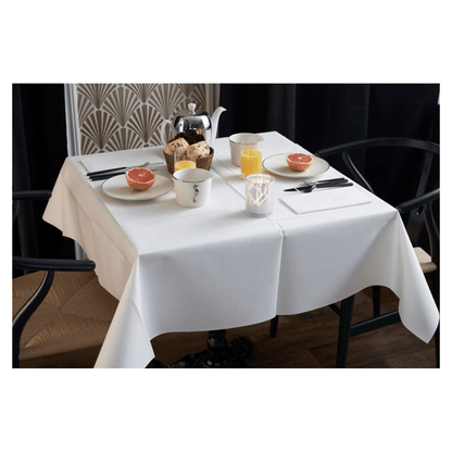 La table du petit-déjeuner est élégamment dressée avec une nappe blanche, deux chaises et un vase à motif géométrique. Sur la table se trouvent des moitiés de pamplemousse, des pâtisseries, du thé et du jus d'orange. Une serviette Duni Dunilin® (48x48 cm) de Duni GmbH ajoute du charme et de la fonctionnalité à l'ensemble matinal d'ambiance.