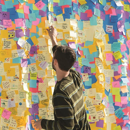 Une personne en chemise rayée se tient devant un mur avec des messages et dessins manuscrits et attrape un cube de notes Post-it® Ultra-Couleurs de 3M Deutschland GmbH (76x76 mm, 450 feuilles, 100% PEFC), pour le placer parmi les notes colorées.