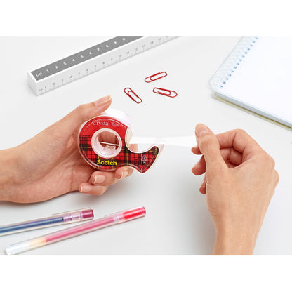 Eine Person zieht Scotch® Crystal Klebeband (19 mm x 7,5 m) aus dem roten Handabroller der 3M Deutschland GmbH auf einem weißen Schreibtisch mit roten Büroklammern, Stiften, einem Lineal und einem Spiralnotizbuch.