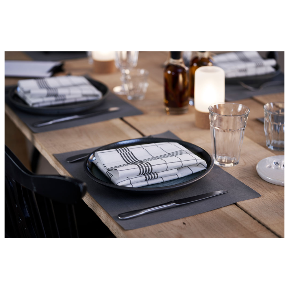 Un ensemble de table à manger composé d'assiettes en Linnea Black de Duni GmbH et de serviettes à carreaux. En arrière-plan, trois bouteilles en verre se tiennent à côté de bougies allumées. Des verres à eau et des couverts sont élégamment présentés sur un set de table Duni Dunicel® 30 x 40 cm, mettant en valeur l'élégance simple de la table en bois clair.