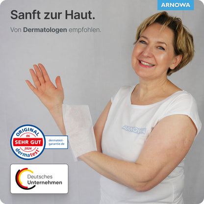Une femme d'âge moyen en chemise blanche sourit en tenant un gant de toilette jetable ARNOMED en molleton (75g/m²) sur son avant-bras. L'image montre du texte en allemand, les logos de Dermatest et de ARNOWA GmbH et met en avant les avis et recommandations sur le produit.