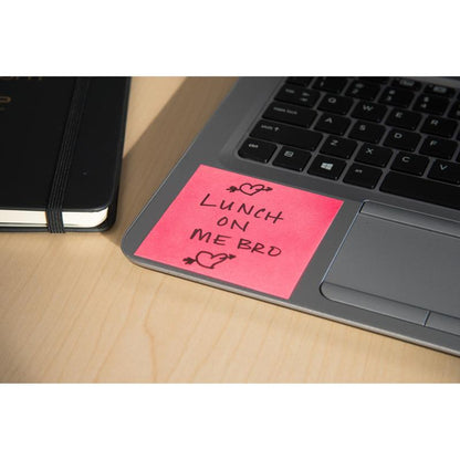Une note Z Super Sticky rose Post-it® (76x76mm) de la société 3M Allemagne avec l'inscription "Lunch on me bro" en encre noire et deux cœurs est posée sur un clavier d'ordinateur portable à côté d'un carnet noir, le tout sur une table en bois clair.