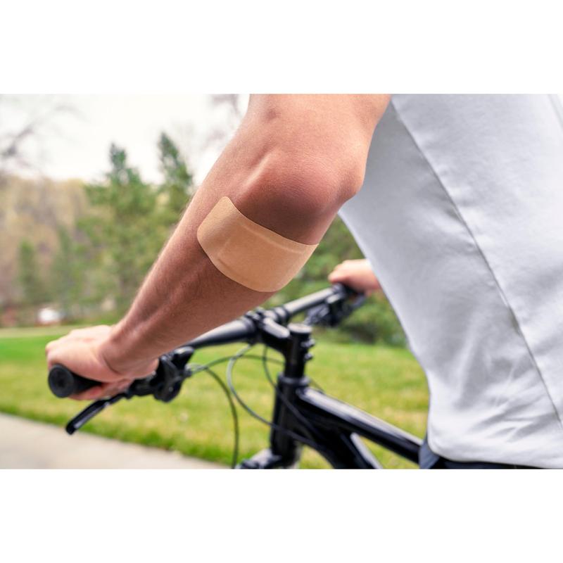 Une personne portant une chemise blanche et faisant du vélo en extérieur a un pansement Nexcare™ Ultra Stretch MAXI Comfort Flexible (50 mm x 101 mm) de 3M Deutschland GmbH sur le bras nu; en arrière-plan, on voit de l'herbe verte et des arbres.