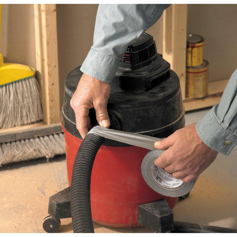 Une personne portant une chemise à manches longues utilise le ruban adhésif Scotch® Universal 2904 (25 m x 48 mm) de 3M Deutschland GmbH pour fixer le tuyau d'un aspirateur d'atelier rouge et noir. En arrière-plan, on voit un balai et des pots de peinture.