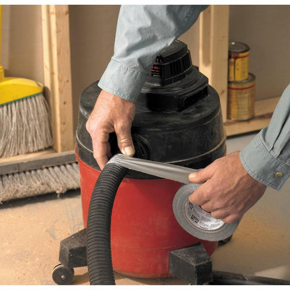 Une personne portant une chemise à manches longues utilise le ruban adhésif Scotch® Universal 2904 (25 m x 48 mm) de 3M Deutschland GmbH pour fixer le tuyau d'un aspirateur d'atelier rouge et noir. En arrière-plan, on voit un balai et des pots de peinture.