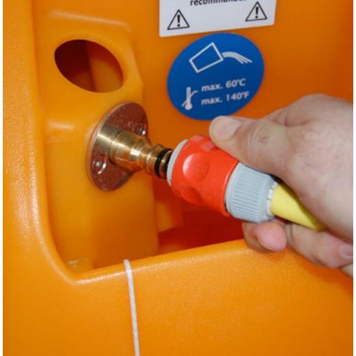 Une main connecte un tuyau avec un raccord rouge et gris à un raccord métallique sur un TASKI swingo 2100 ?icro AquaStop orange de Diversey Deutschland GmbH & Co. OHG. Un autocollant bleu à proximité indique une température maximale de 60°C/140°F.
