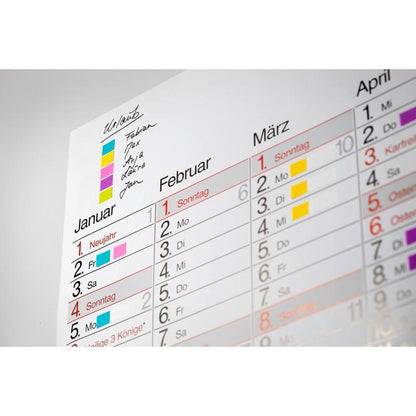 Gros plan d'un calendrier mural de janvier à mars avec des Post-it® Index Signets colorés de 3M Deutschland GmbH (Mini, 11,9 x 43,2 mm) comme marqueurs codés par couleur pour les rendez-vous ou activités manuscrits de chaque personne.