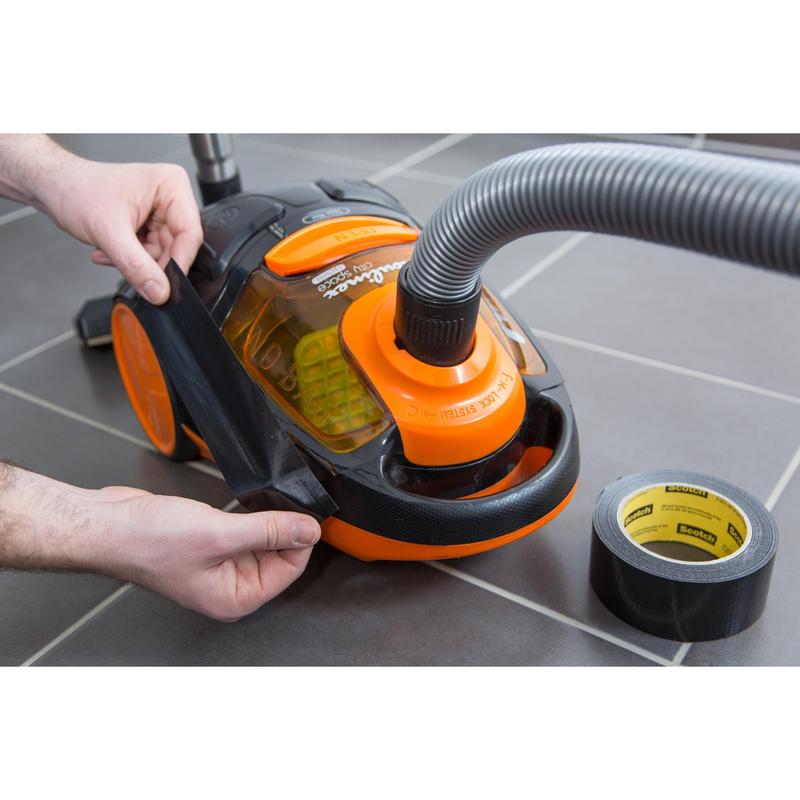 Une personne enroule du ruban adhésif Scotch® Universal 2904 (25 m x 48 mm) de 3M Deutschland GmbH autour du support de roue d'un aspirateur orange et noir sur un sol carrelé gris, avec le tuyau de l'aspirateur fixé et un rouleau de ruban à proximité.