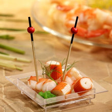 Sur un plat carré transparent se trouvent des imitations de crabe, des crevettes, une garniture verte et deux PAPSTAR 250 picots à amuse-bouches de la PAPSTAR Une GmbH ornée de perles rouges. À l'arrière-plan, on aperçoit des crevettes floues posées sur une assiette.