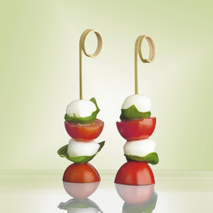 Deux PAPSTAR 100 piques à finger food « pure » de PAPSTAR GmbH en plastique de haute qualité tiennent des boules de mozzarella, du basilic et des tomates cerises, décorées devant un vert doux – idéal pour les fêtes et événements.