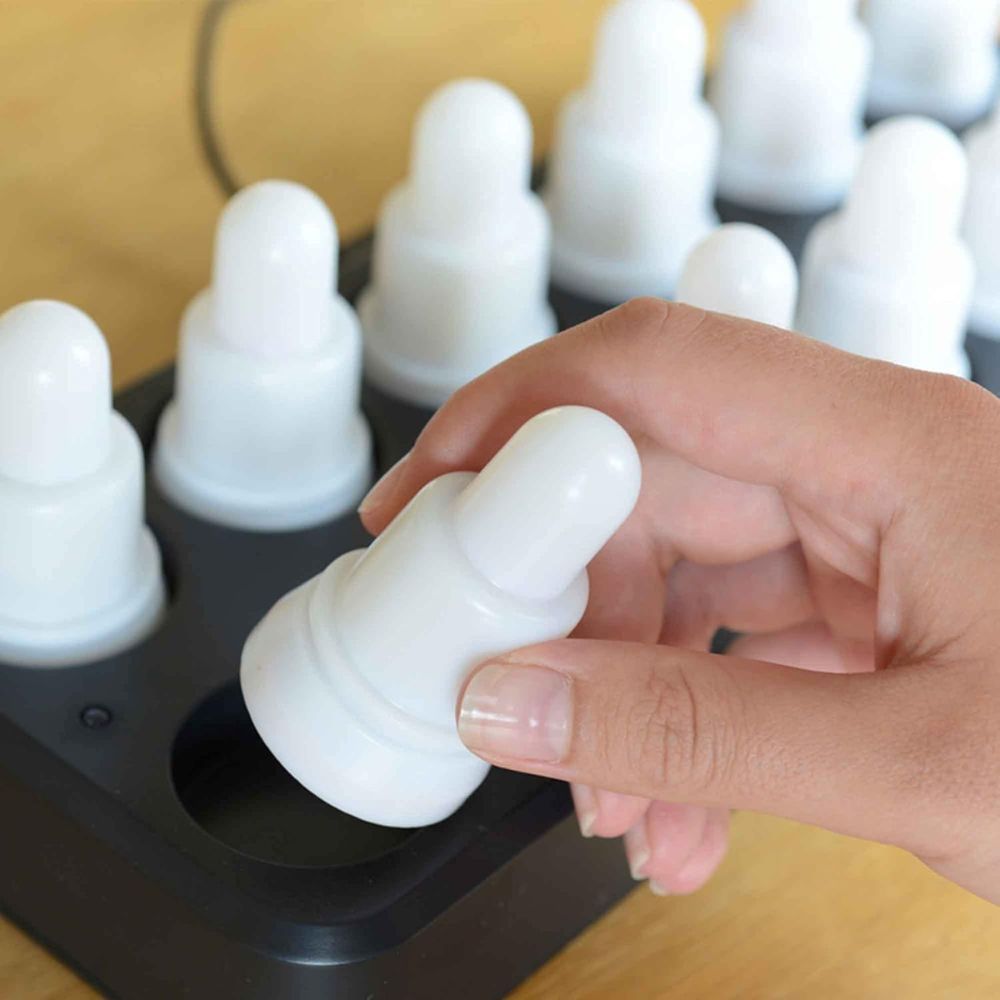 Une main place ou retire une bougie LED ronde blanche du plateau noir du set LED Duni GmbH, Blanc chaud & Multicolore - 12 pièces de lampes à changement de couleur; chaque bougie rechargeable identique repose sur une base en bois.