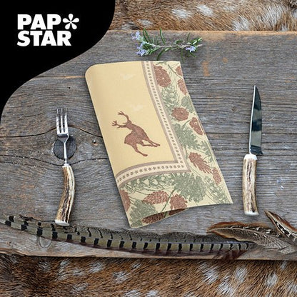 Une serviette de la PAPSTAR GmbH ROYAL Collection (40x40 cm, 1/4 plié) avec motif de cerf et de pommes de pin repose sur une table rustique en bois avec des couverts ressemblant à des bois, des plumes et des herbes. Dans le coin supérieur gauche se trouve le logo PAPSTAR.