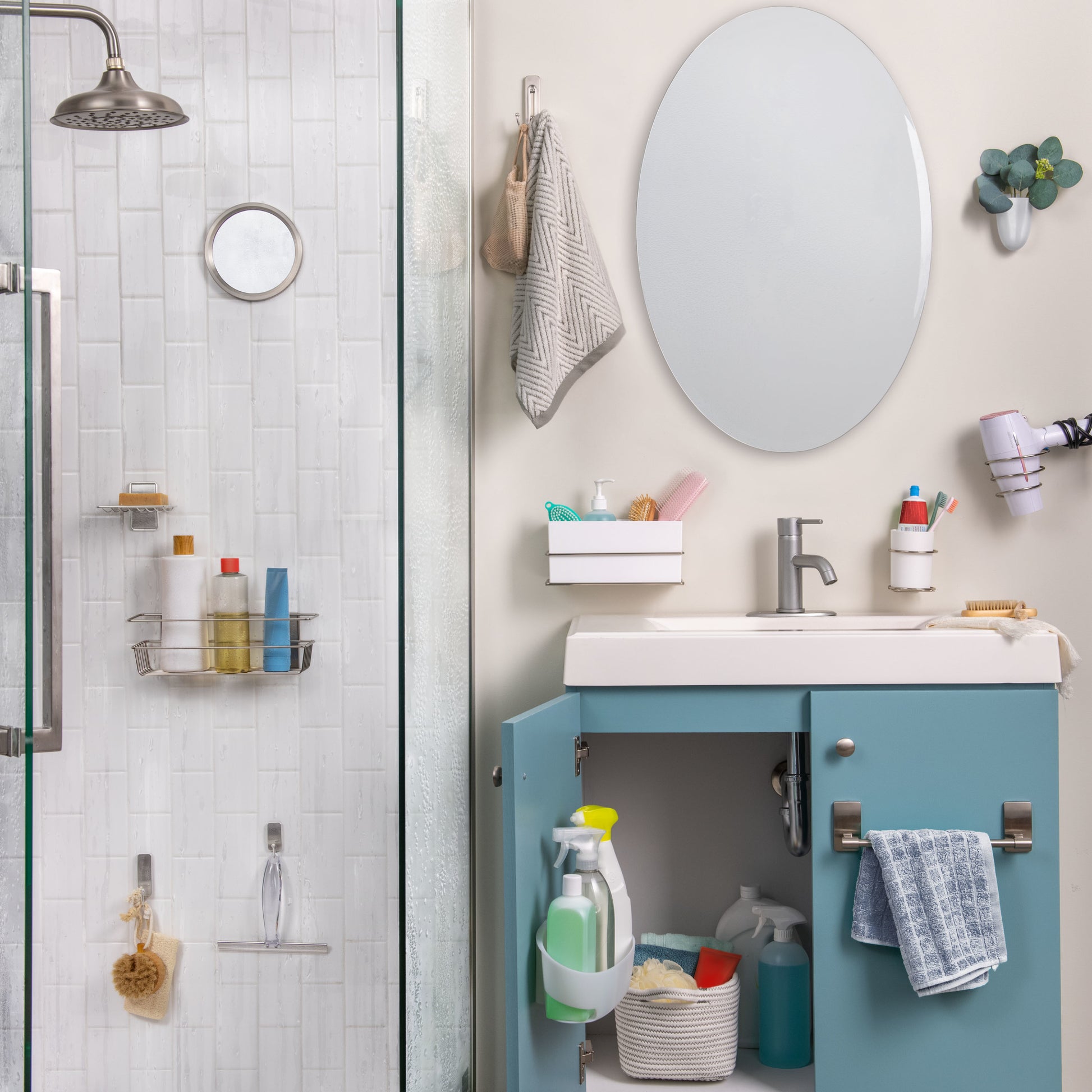 Ein Badezimmer mit einer Glasdusche auf der linken Seite, einem blauen Waschtisch, einem weißen Waschbecken, einem ovalen Spiegel und organisierten Toilettenartikeln. Handtücher hängen ordentlich am 3M Command™ Großer Badezimmer-Doppelhaken Nickel satiniert von 3M Deutschland GmbH nebenan.