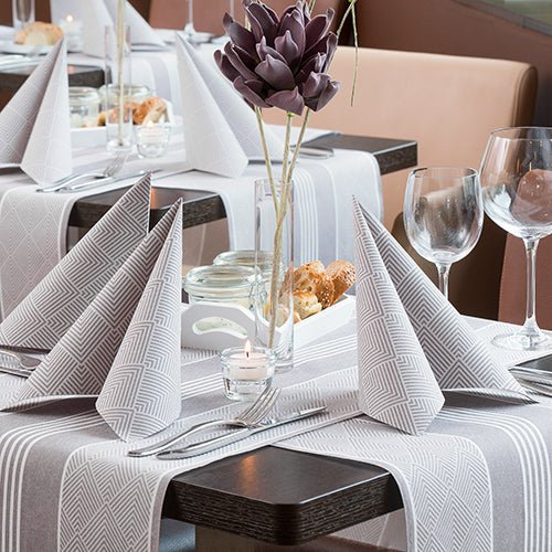 Une table de restaurant dressée avec PAPSTAR GmbH 50 serviettes "ROYAL Collection" 1/4-pli, 40 x 40 cm, marron, pliées en triangles, avec des verres à vin, des couverts, un panier à pain, une pièce maîtresse en fleurs artificielles violettes et une bougie chauffe-plat allumée.
