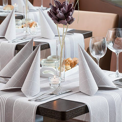 Une table de restaurant dressée avec PAPSTAR GmbH 50 serviettes "ROYAL Collection" 1/4-pli, 40 x 40 cm, marron, pliées en triangles, avec des verres à vin, des couverts, un panier à pain, une pièce maîtresse en fleurs artificielles violettes et une bougie chauffe-plat allumée.