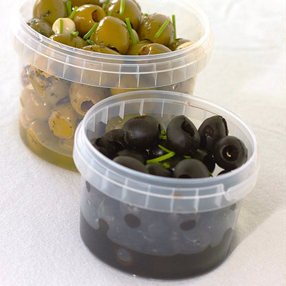 Dans deux gobelets transparents Starpak 30 de 770 ml (PP, Ø 11,8 cm · 11,3 cm) avec couvercle d'authenticité, des olives sont disposées sur un fond blanc - dans le gobelet arrière, des olives vertes aux herbes et à l'avant, des olives noires avec garniture d'herbes.