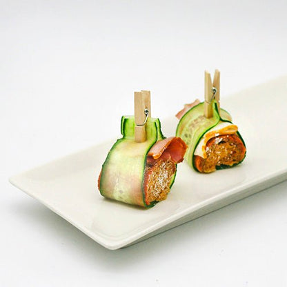 Deux amuse-gueules enveloppés de tranches de concombre sont sécurisés avec des PAPSTAR mini-pinces en bois (3,5 cm) de PAPSTAR GmbH, idéal pour les projets de bricolage, et servis sur un plateau en carton (100 pièces) - parfait pour des plateaux de fête créatifs.