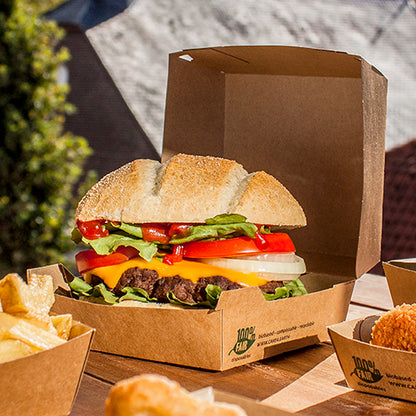 Un cheeseburger avec de la laitue, de la tomate, de l'oignon et du fromage cheddar est placé en plein air dans une boîte à burger en carton PAPSTAR GmbH (9x18,5x18,5 cm, "100% Fair", 25 pièces). Les frites et d'autres accompagnements sont servis séparément à côté.