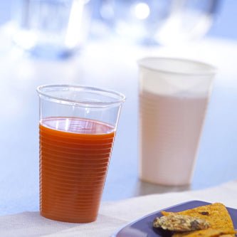 Un gobelet Starpak 25, PP 0,2 l (Ø 7,03 cm · 9,9 cm), rempli de jus d'orange, est au premier plan à côté d'une tasse blanche avec une boisson claire ; en bas à droite, une assiette bleue avec de la nourriture est visible – idéal pour les déplacements.