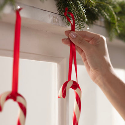 Eine Hand hängt eine Zuckerstange mit einem roten Band an einen Command™ Mini-Haken, transparent, mit transparenten Strips (3M Deutschland GmbH), der unter einem mit immergrünen Zweigen dekorierten Regal angebracht ist. Eine weitere Zuckerstange hängt bereits an dem Haken daneben.