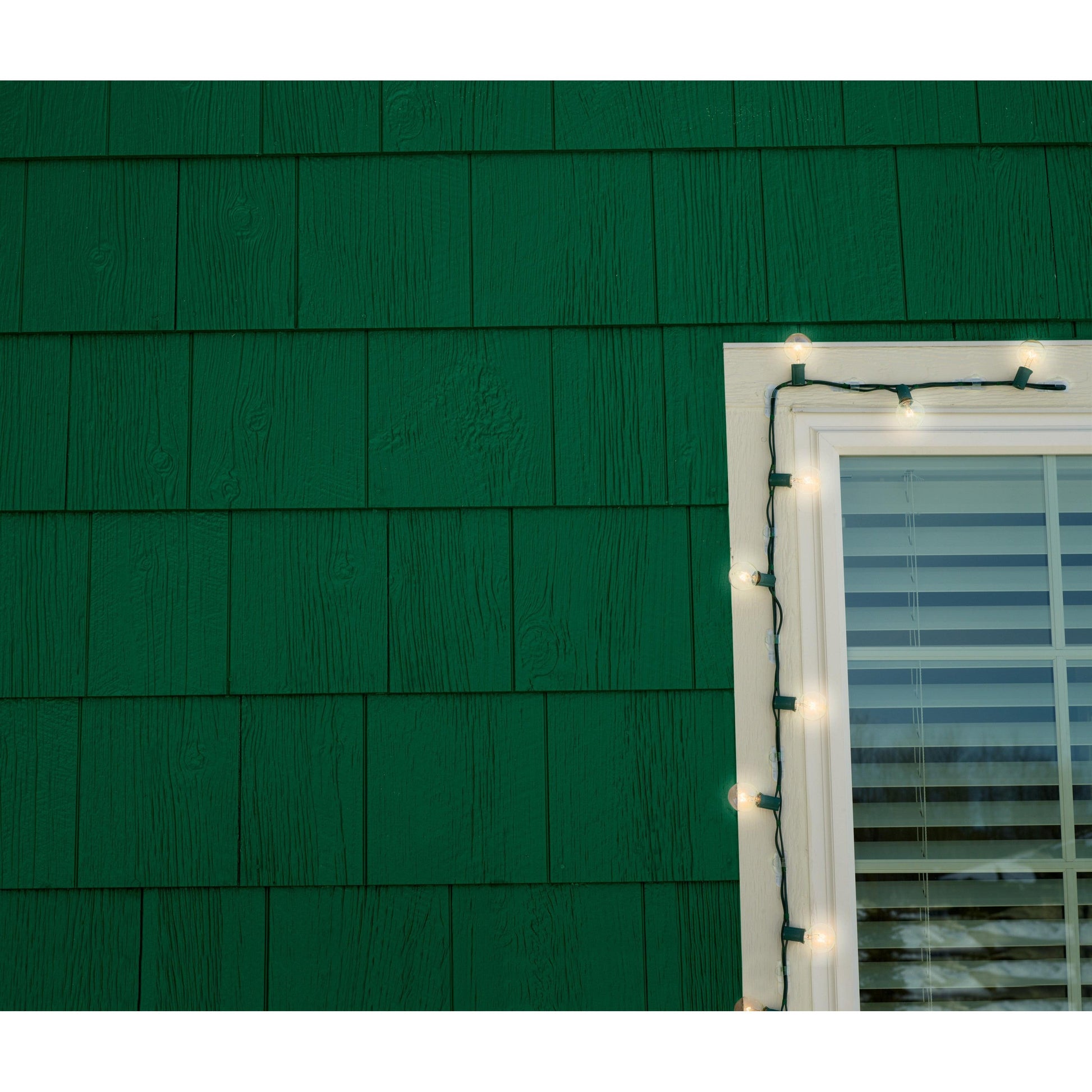 In einem Haus mit grüner Fassade und weiß umrahmten Fenstern werden 3M Deutschland GmbH Command™ Outdoor Lichter-Clips (17017CLR-AWPU, 8 Clips + 10 Streifen) verwendet, um eine weiße Lichterkette senkrecht neben dem Fenster aufzuhängen.