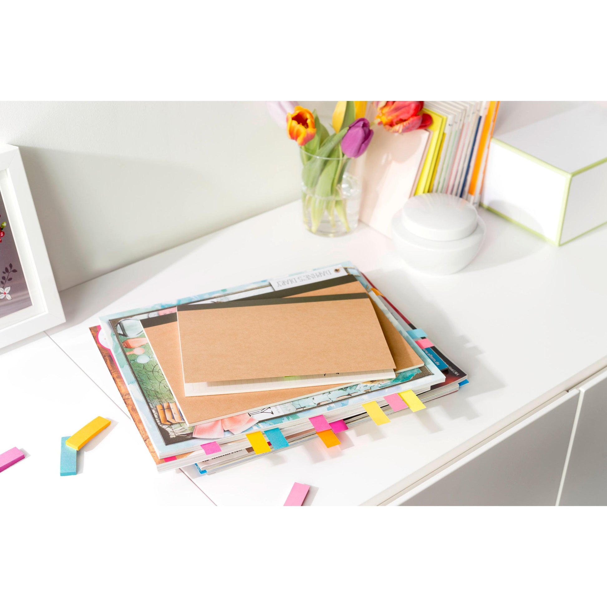 Ein Stapel Bücher und Notizbücher mit bunten Post-it® Page Markern (15x50mm, 6+3 Promo-Pack) der 3M Deutschland GmbH liegt auf einem weißen Schreibtisch neben Tulpen, Kunst, einem weißen Container und anderen Büchern und Schachteln.