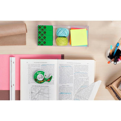 Un bureau avec un livre ouvert, des classeurs, des crayons de couleur dans un support, des notes adhésives, deux boules de ficelle, plusieurs gommes vertes, un rouleau de papier brun et un paquet de ruban adhésif invisible Scotch® Magic™ (8 rouleaux, 19 mm x 33 m) de 3M.