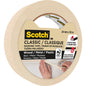 Un rouleau de ruban de masquage 3M Scotch® Classic, 24 mm x 50 m, 70% PEFC (SGSCH-COC-110078), idéal pour les travaux de peinture et de rénovation sur bois, métal, plastique. La forte adhérence dure jusqu'à deux jours. L'image montre une main appliquant le ruban.