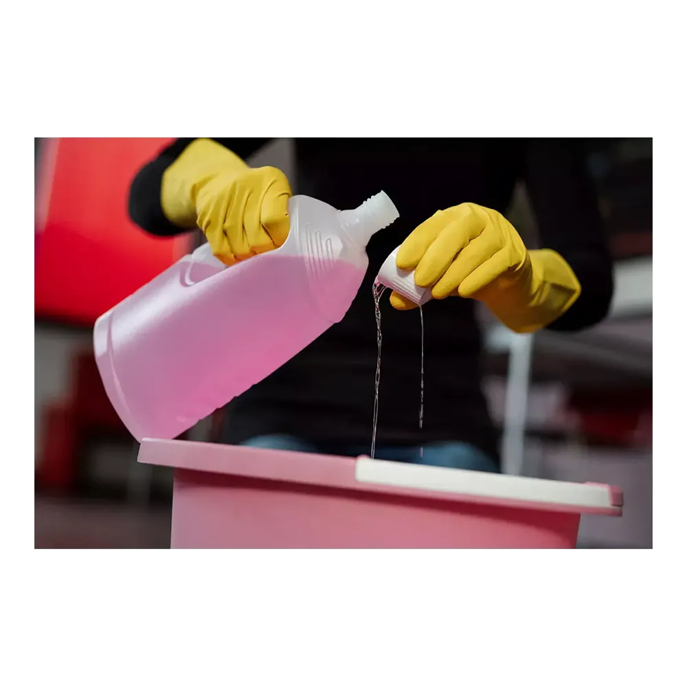 Une personne portant des gants de protection chimique AMPri SolidSafety ChemL de AMPri Handelsgesellschaft mbH verse un liquide d'une bouteille en plastique transparente dans un petit couvercle au-dessus d'un récipient rose. Le liquide, peut-être une solution de nettoyage AMPri, se détache sur le fond flou.