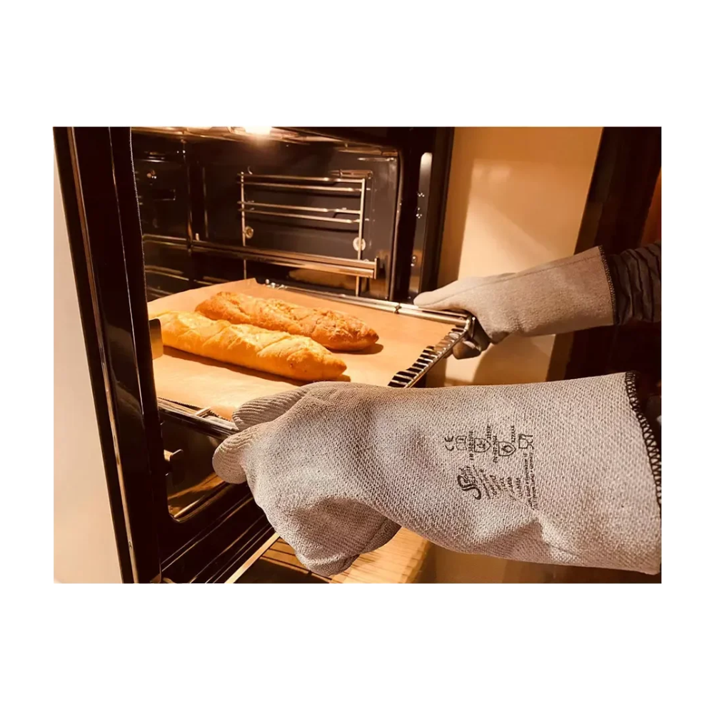 Une personne avec les gants de protection contre la chaleur AMPri SolidSafety L - XXL de la société commerciale AMPri retire prudemment une plaque de cuisson du four. Sur la plaque se trouvent deux miches de pain, tandis que l'éclairage chaud de la cuisine met en valeur à la fois le four ouvert et la protection maximale contre la chaleur de ces gants de protection.