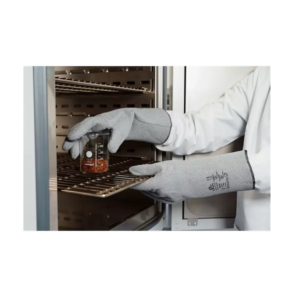 Une personne porte les gants de protection contre la chaleur AMPri SolidSafety L - XXL de la société commerciale AMPri et une blouse de laboratoire et place prudemment un bécher en verre avec un liquide rougeâtre sur un support métallique dans un four industriel. L'accent est mis sur les mains et le bécher, ce qui souligne la protection maximale contre la chaleur, tandis que les étagères à l'intérieur de l'appareil sont visibles.