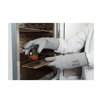 Une personne porte les gants de protection contre la chaleur AMPri SolidSafety L - XXL de la société commerciale AMPri et une blouse de laboratoire et place prudemment un bécher en verre avec un liquide rougeâtre sur un support métallique dans un four industriel. L'accent est mis sur les mains et le bécher, ce qui souligne la protection maximale contre la chaleur, tandis que les étagères à l'intérieur de l'appareil sont visibles.