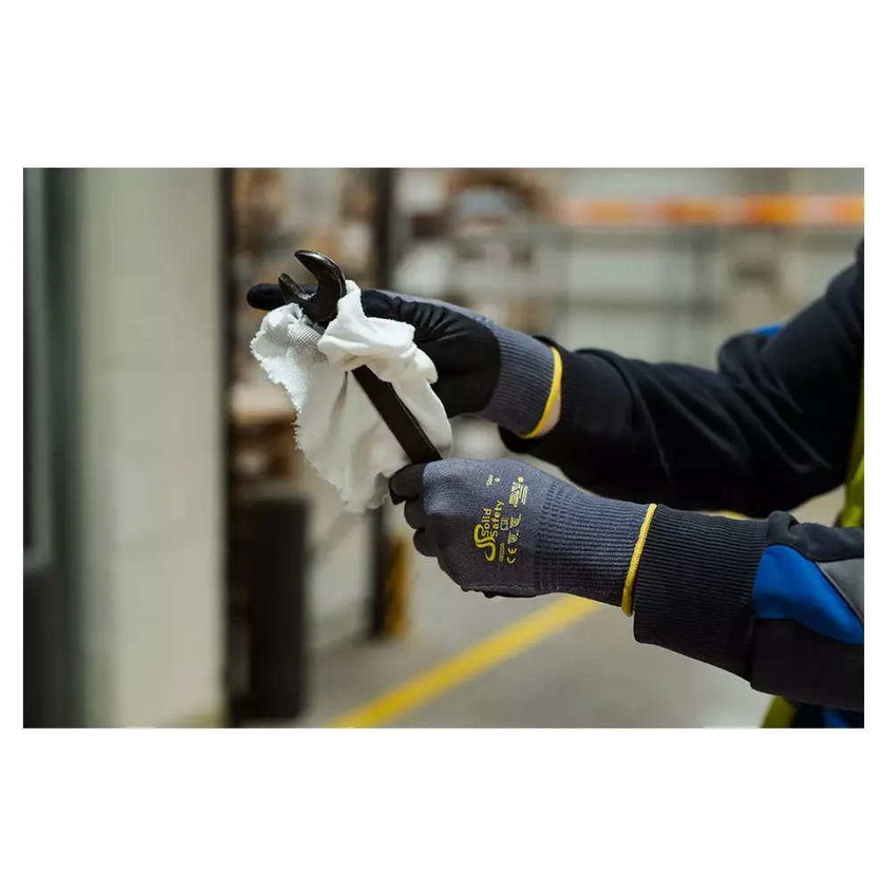 Une personne avec des gants de montage ultralégers AMPri SolidSafety Tough Air de la société commerciale AMPri Handelsgesellschaft mbH nettoie soigneusement une clé à molette avec un chiffon blanc à l'intérieur. Les gants gris, reconnus pour leur niveau de résistance à l'abrasion de 4, se détachent des étagères et équipements industriels flous en arrière-plan.