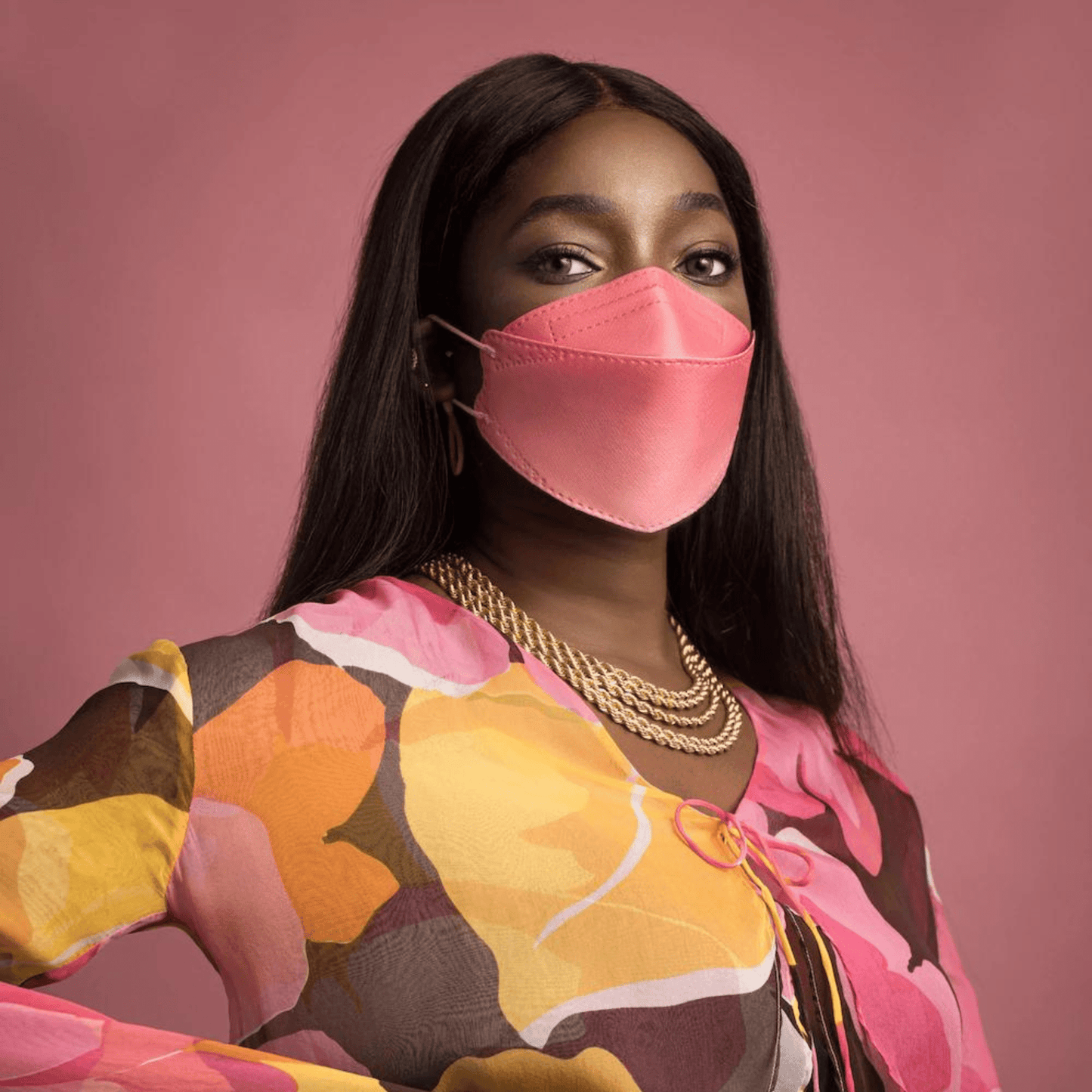 Une personne porte un masque facial de designer Altruan filtrant les particules (norme FFP2) dans une teinte rose élégante et un haut coloré avec un motif abstrait devant un fond rose. Elle a de longs cheveux foncés et porte plusieurs colliers en or.