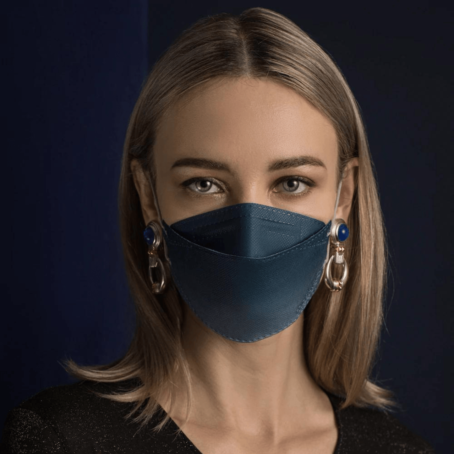 Une personne aux longs cheveux porte un masque facial de designer Altruan filtrant les particules en bleu, offrant une protection efficace selon la norme FFP2. Elle le combine avec de grandes boucles d'oreilles décoratives devant un fond sombre et porte une chemise noire.