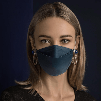 Une personne aux longs cheveux porte un masque facial de designer Altruan filtrant les particules en bleu, offrant une protection efficace selon la norme FFP2. Elle le combine avec de grandes boucles d'oreilles décoratives devant un fond sombre et porte une chemise noire.