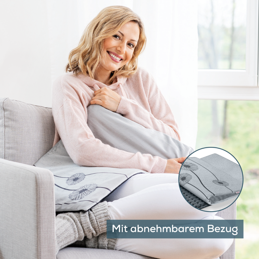 Une femme est assise sur un canapé gris clair près d'une fenêtre et tient le Beurer coussin chauffant HK 125 XXL Cosy Grey de Beurer GmbH dans la main. Elle sourit et porte un sweat à capuche rose. Il y a un gros plan du coussin, montrant sa housse amovible et ses fonctions de chauffage avec 6 niveaux de chaleur. Le texte allemand indique « Avec housse amovible. »