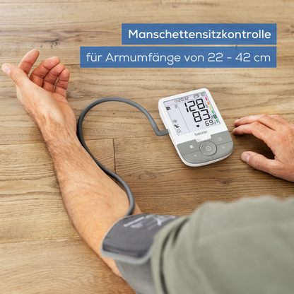 Un utilisateur travaille avec le Beurer GmbH article discontinué : tensiomètre à bras BM 53 sur une surface en bois clair. L'écran affiche "Contrôle du positionnement du brassard pour des circonférences de bras de 22 à 42 cm" et indique la détection d'arythmie et la prévention des AVC.