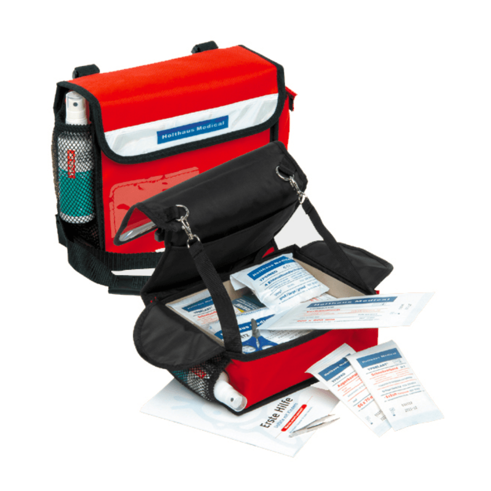 Un sac médical rouge Holthaus pour la maternelle de Holthaus Medical GmbH & Co. KG est ouvert et montre une sélection d'articles de premiers secours, y compris des bandages, des pansements, un livret de premiers secours et une petite bouteille. Le sac a un rabat noir et des poches latérales en maille. Il est idéal comme trousse de premiers secours dans des environnements tels que les jardins d'enfants.
