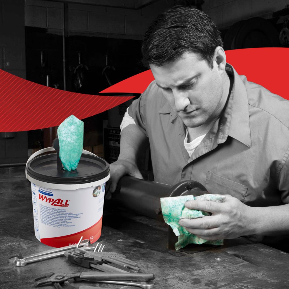 Un homme en tenue de travail nettoie un tuyau métallique avec une lingette verte WypAll® de Kimberly-Clark GmbH. Un carton (6 seaux distributeurs) de lingettes est posé parmi les outils. La photo est principalement en noir et blanc, à l'exception des lingettes et de l'étiquette.