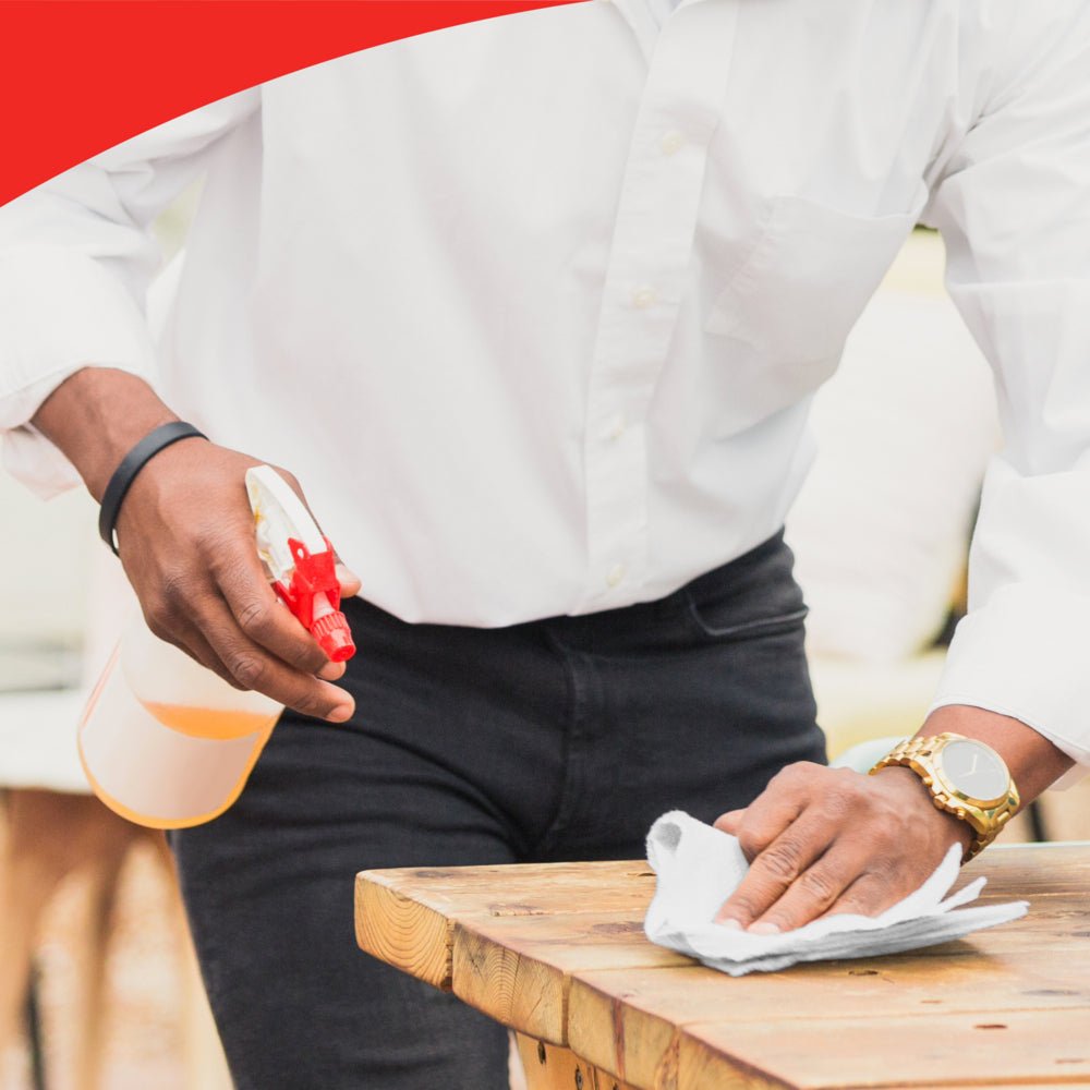 Une personne en chemise blanche et pantalon noir essuie une table en bois avec un grand rouleau WypAll® X50 General Clean de Kimberly-Clark GmbH (1 rouleau x 1100 feuilles). Seul son torse et ses mains sont visibles. L'image a un bord ondulé rouge en haut.