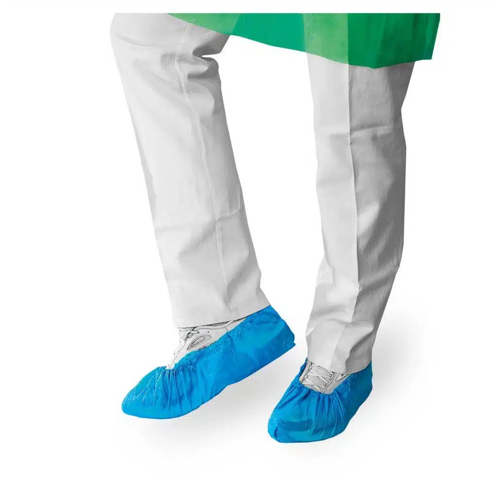 Une personne en blouse de laboratoire blanche et en couvre-chaussures bleus jetables de Meditrade GmbH se tient devant un fond blanc, avec seulement ses mollets visibles.