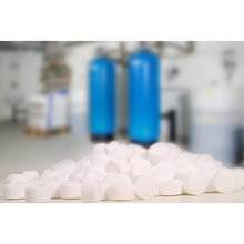 Im Vordergrund fällt ein Stapel Regenit Siedesalztabletten, gemäß EN 973 Typ, der K+S Minerals and Agriculture GmbH auf, während der unscharfe Hintergrund zwei große blaue Wasserenthärtungsanlagen erkennen lässt. Die industrielle Kulisse lässt auf einen präzisen Nutzraum schließen, der den Normen EN 973 Typ A entspricht.