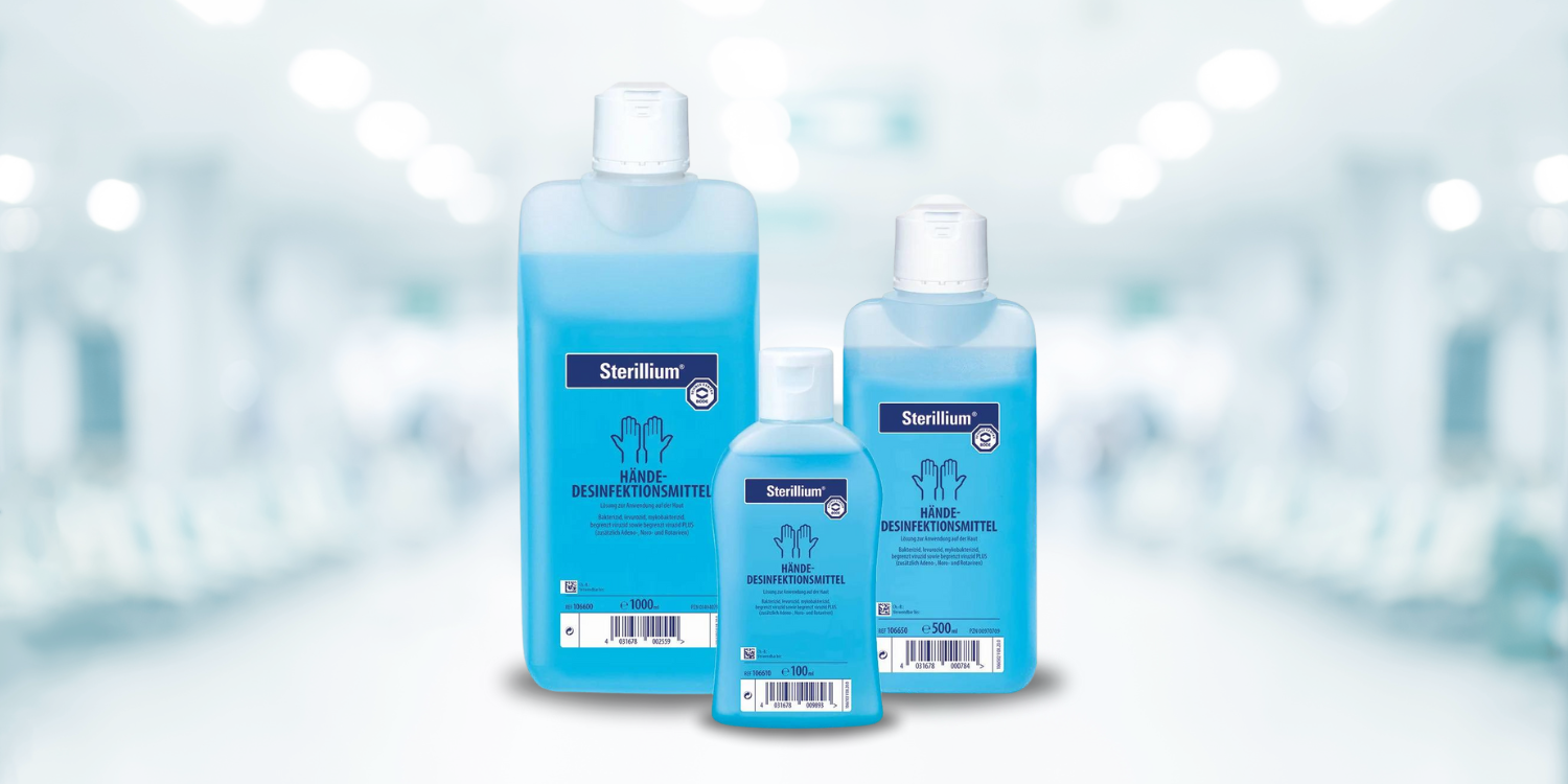 Das Bild zeigt drei Flaschen mit Desinfektionsmittel der Marke Sterillium in unterschiedlichen Größen, die in einer Reihe auf einer weißen Oberfläche stehen. Die Flaschen sind blau gefüllt und haben weiße Verschlüsse. Im Hintergrund ist ein unscharfer Innenraum, der wie ein Krankenhausflur aussieht, zu sehen. Die Umgebung ist hell beleuchtet, und das Ambiente vermittelt eine saubere und sterile Atmosphäre.