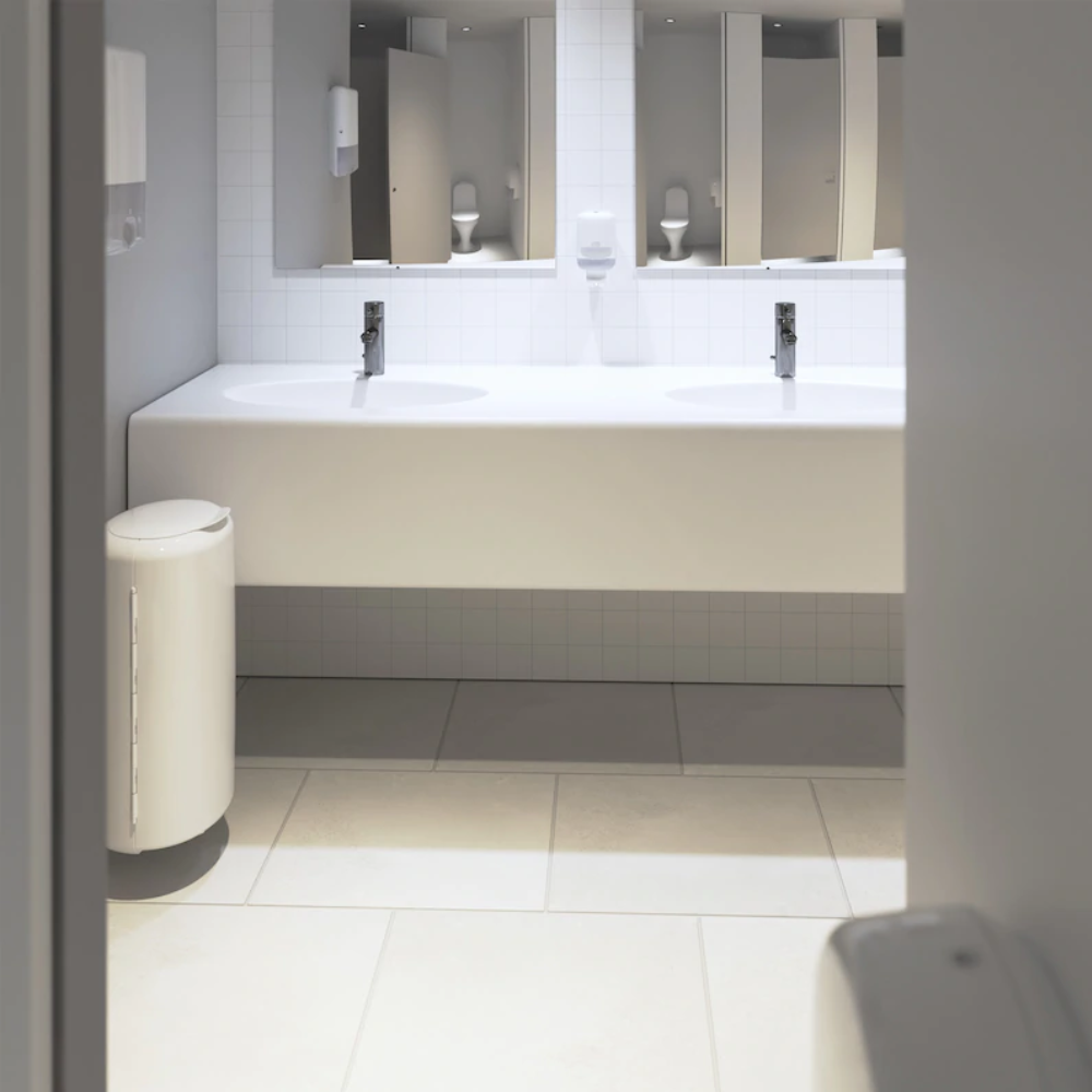 Eine moderne öffentliche Toilette verfügt über zwei weiße Waschbecken mit rechteckigen Spiegeln darüber, umgeben von glatten hellgrauen Fliesen. Ein Tork 561000 Mini Spender für Flüssigseife Elevation S2 von Essity Professional Hygiene Germany GmbH ist neben den Waschbecken angebracht, zusammen mit einem weißen Mülleimer, um für Sauberkeit zu sorgen.
