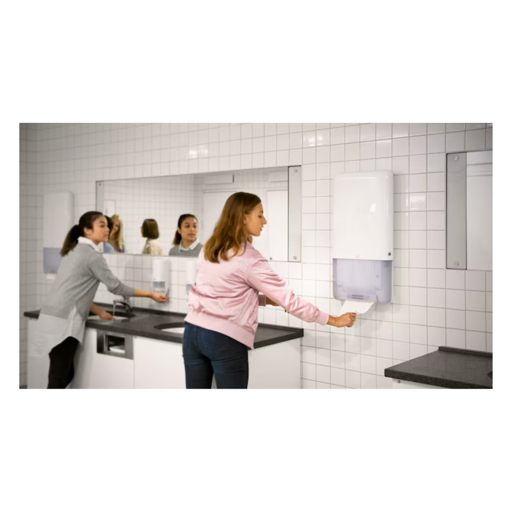 Deux femmes se tiennent dans une salle de bain avec des murs carrelés blancs et de grands miroirs. L'une utilise un lavabo sur le côté gauche, tandis que l'autre tire sans effort une serviette sans fin d'un distributeur Tork PeakServe® 552500 pour serviettes sans fin Elevation H5 de TORK – Essity Professional Hygiene Germany GmbH, situé sur le côté droit. Le plan de travail gris complète l'éclairage lumineux de cette salle de bain très fréquentée.